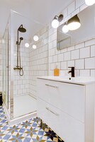 Un baño moderno con paredes de azulejos blancos, piso de diseño geométrico, cabina de ducha de vidrio y tocador con espejo e iluminación.