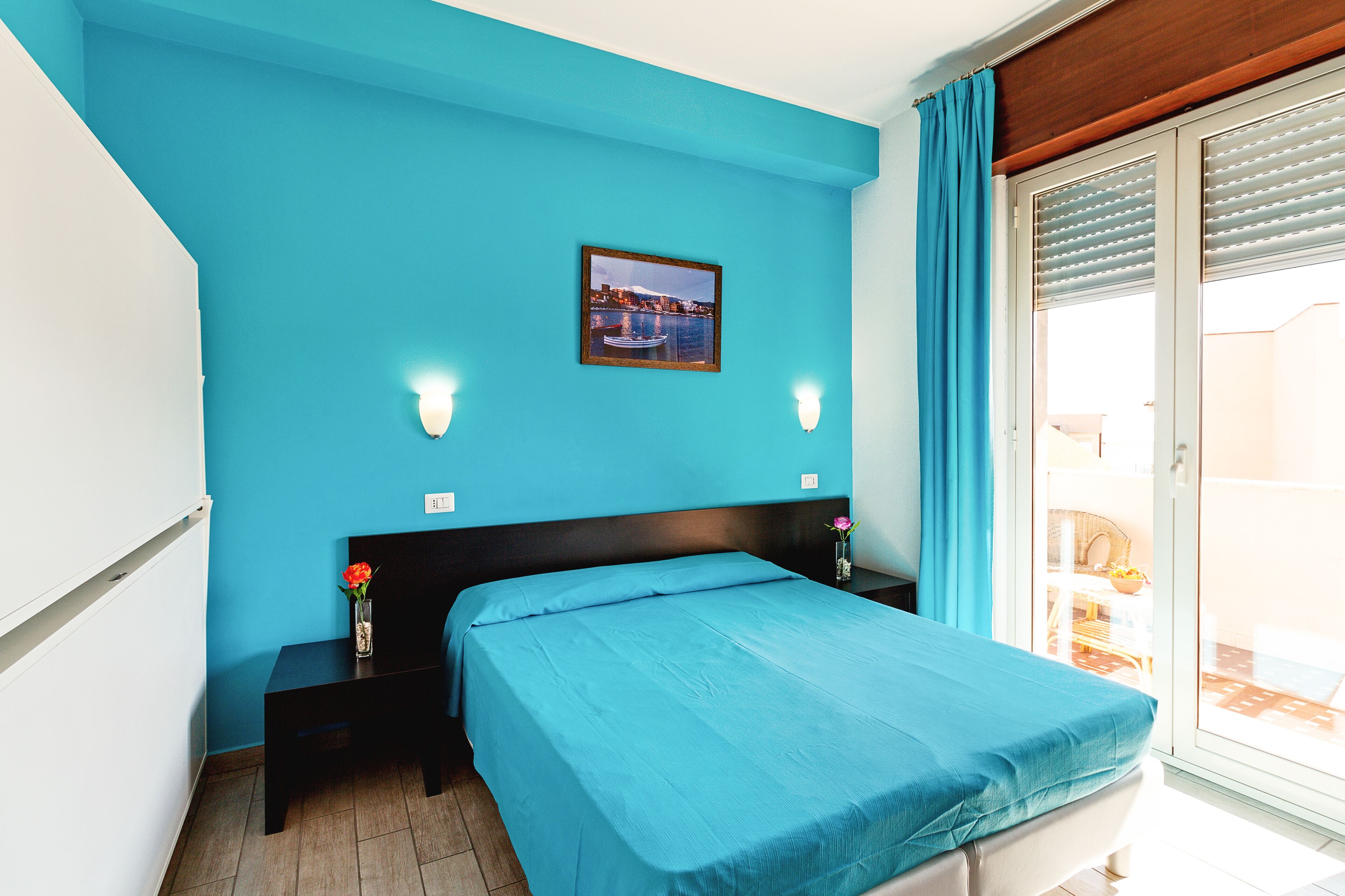 Un dormitorio con pared azul, cama con colcha azul, mesita de noche con un florero y un cuadro en la pared.
