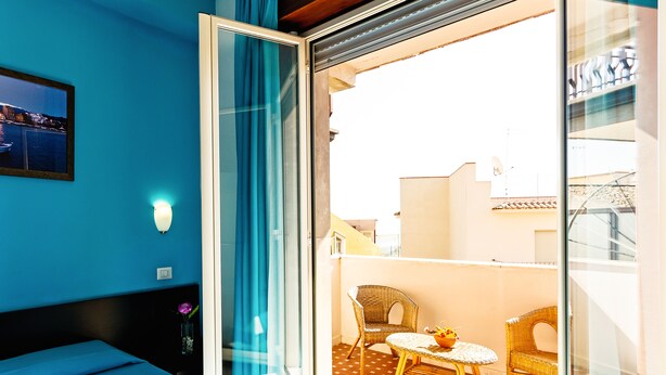 Quarto com cama, janela com cortinas, luminária na parede e vista para uma varanda com mobiliário de vime.
