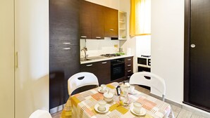 Uma cozinha compacta com armários de madeira escura, bancada branca e uma mesa de jantar para quatro pessoas com toalha de mesa estampada.
