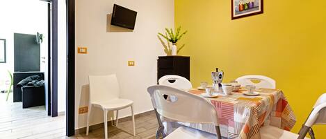 A dining area with a table set for four, white chairs, a yellow wall, a TV mounted on the wall, and a small framed picture.