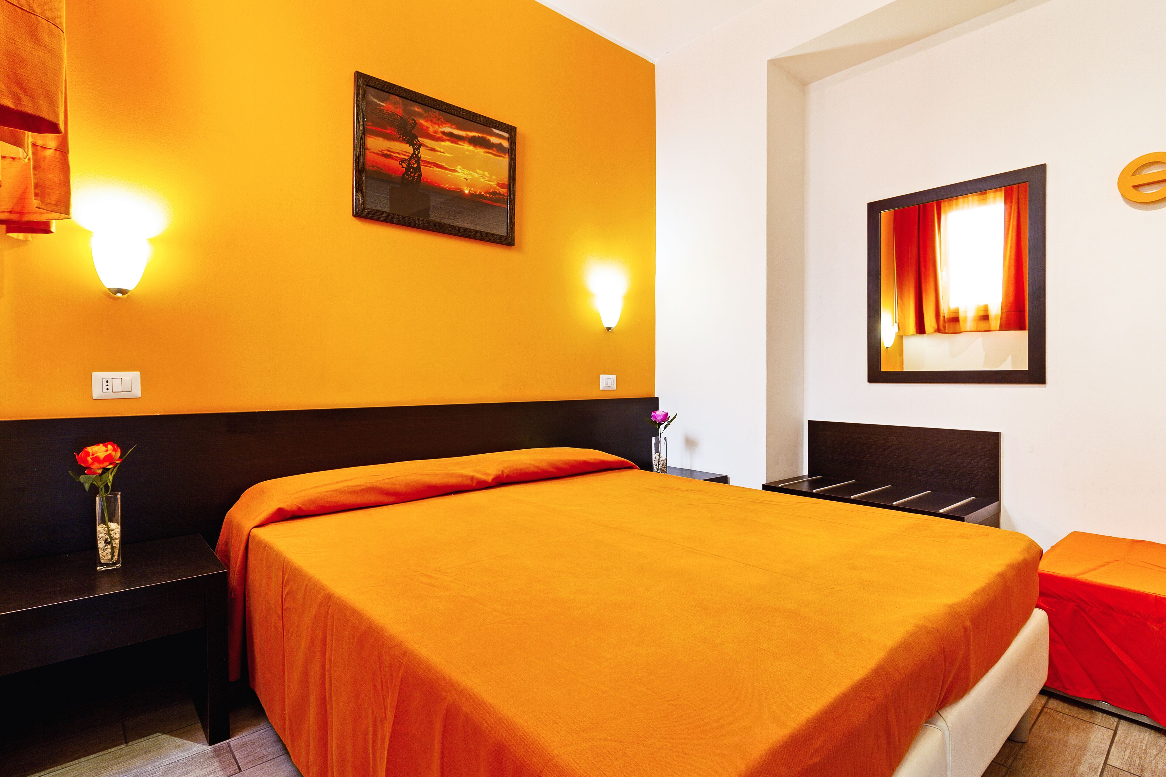 A hotel room with a large bed, two bedside tables, a nightstand, a vase with a flower, and two wall-mounted lights.
