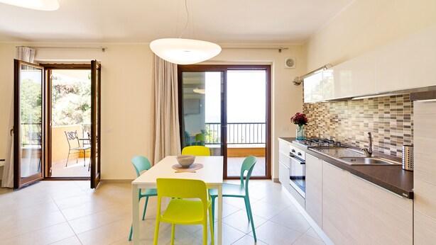 A modern kitchen with a dining area, featuring a large window, sliding glass doors, and a balcony.
