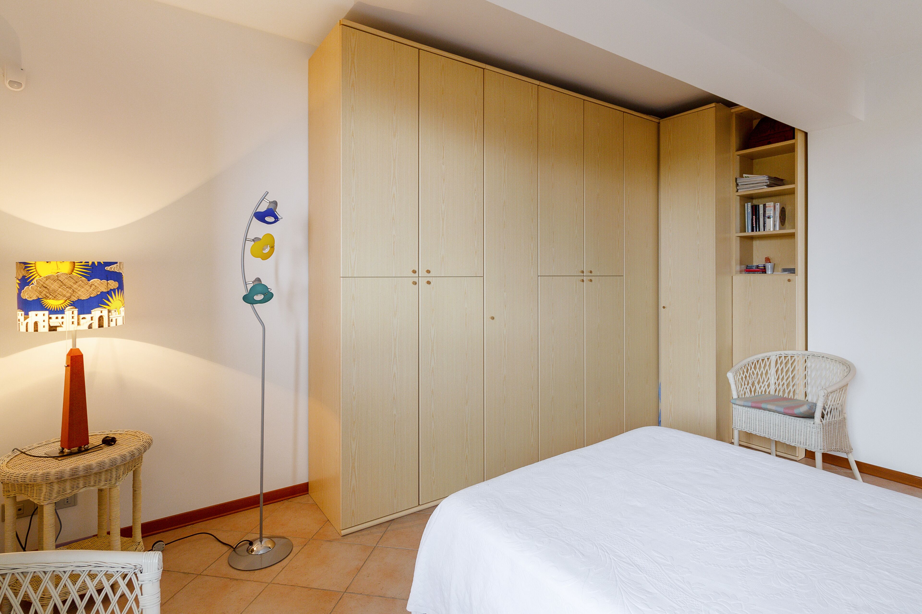 A bedroom with a bed, a wardrobe, a chair, a floor lamp, and a bookshelf.