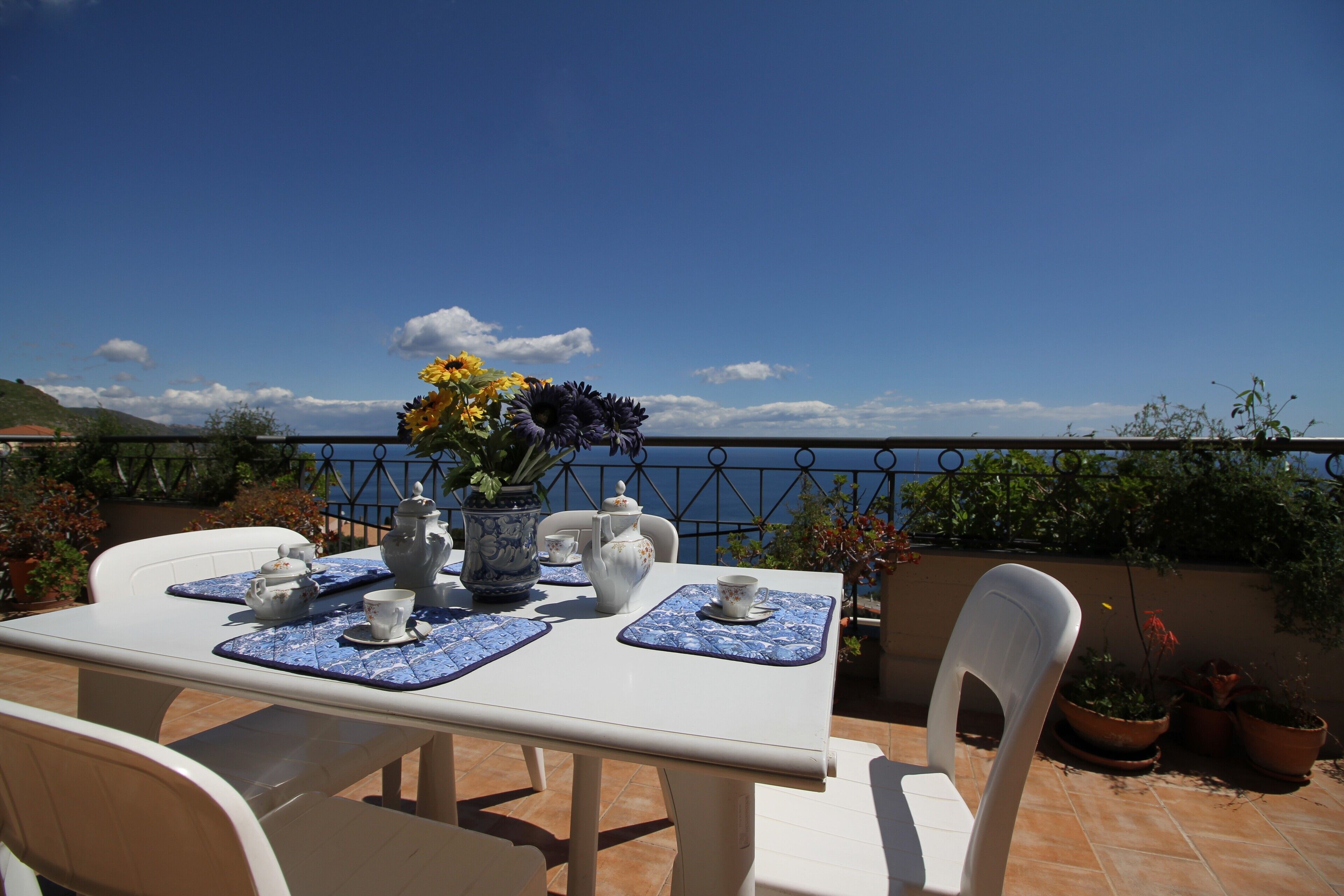 A white table set with blue placemats, a white teapot, cups, and a vase with flowers.
