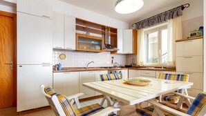 A kitchen with white cabinets, a white dining table with blue and yellow striped chairs, and a window with curtains. - Fontana Vecchia Seaview Apartment  (Taormina)