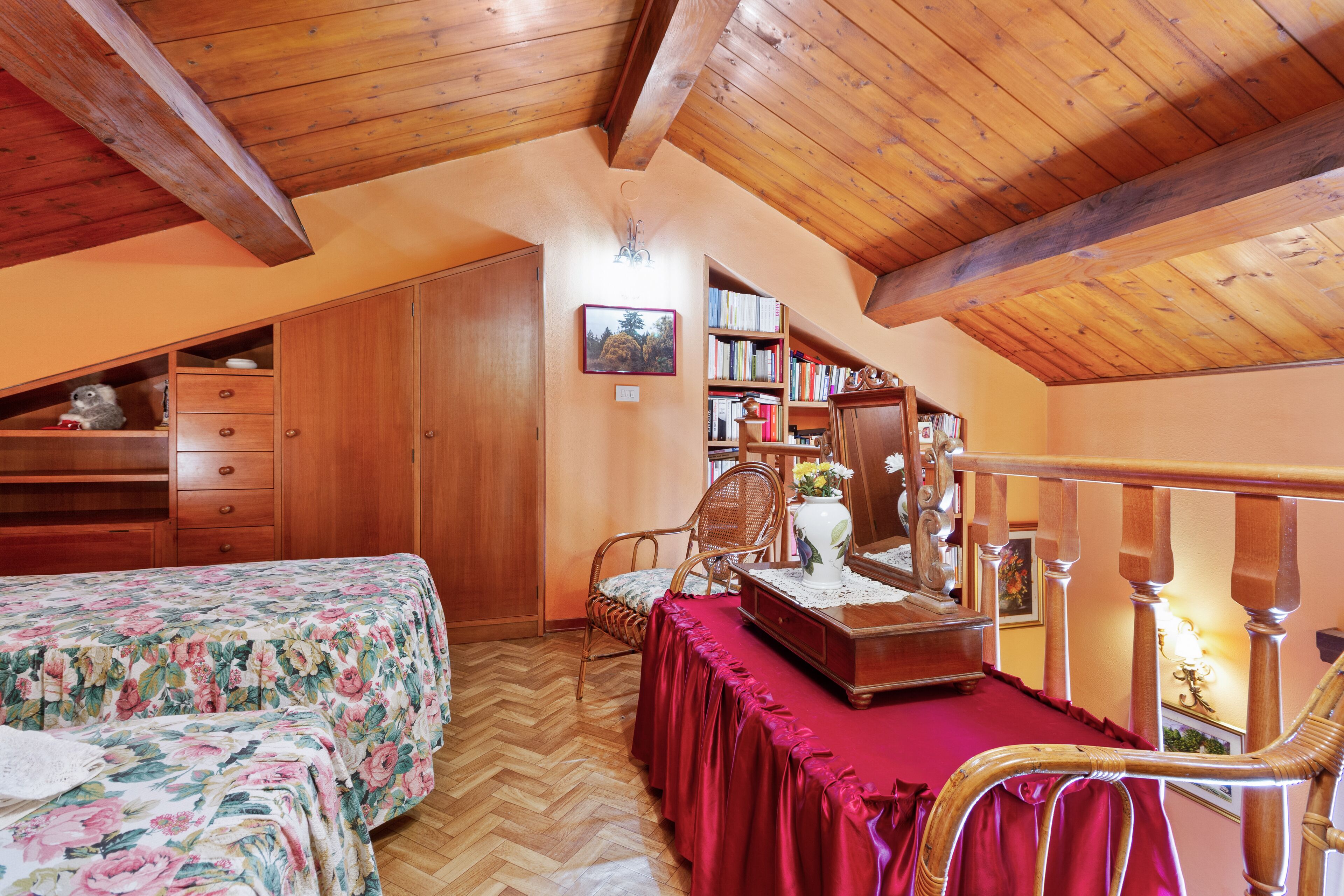A cozy attic bedroom with a wooden ceiling, a bed with floral bedding, a desk with a vase, and bookshelves.