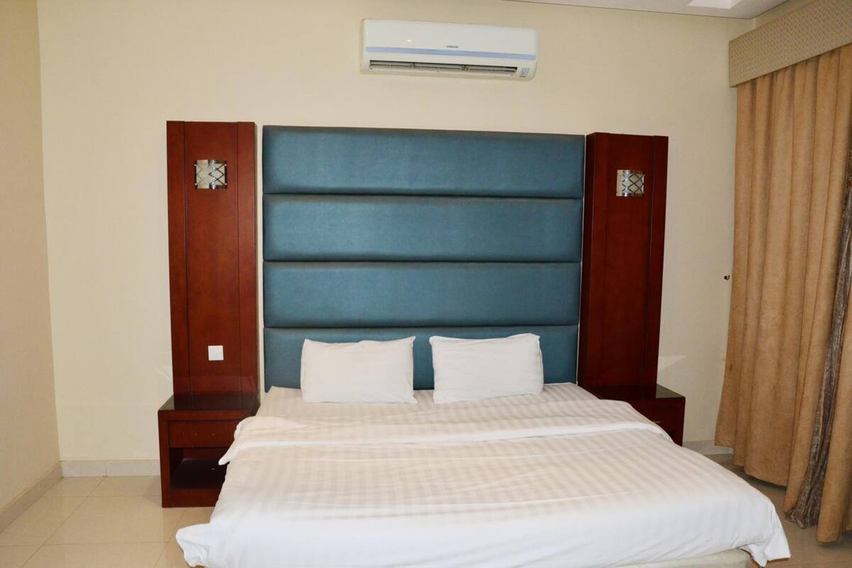 A hotel room with a large bed, a wooden headboard, a wall-mounted air conditioner, and a window with curtains.