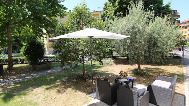 An outdoor seating area with wicker furniture and a white umbrella, surrounded by trees and a pathway. - R&B San Rafèl (Bologna)