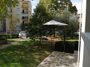 Une cour avec une terrasse en pierre, un parasol blanc et un arbre.