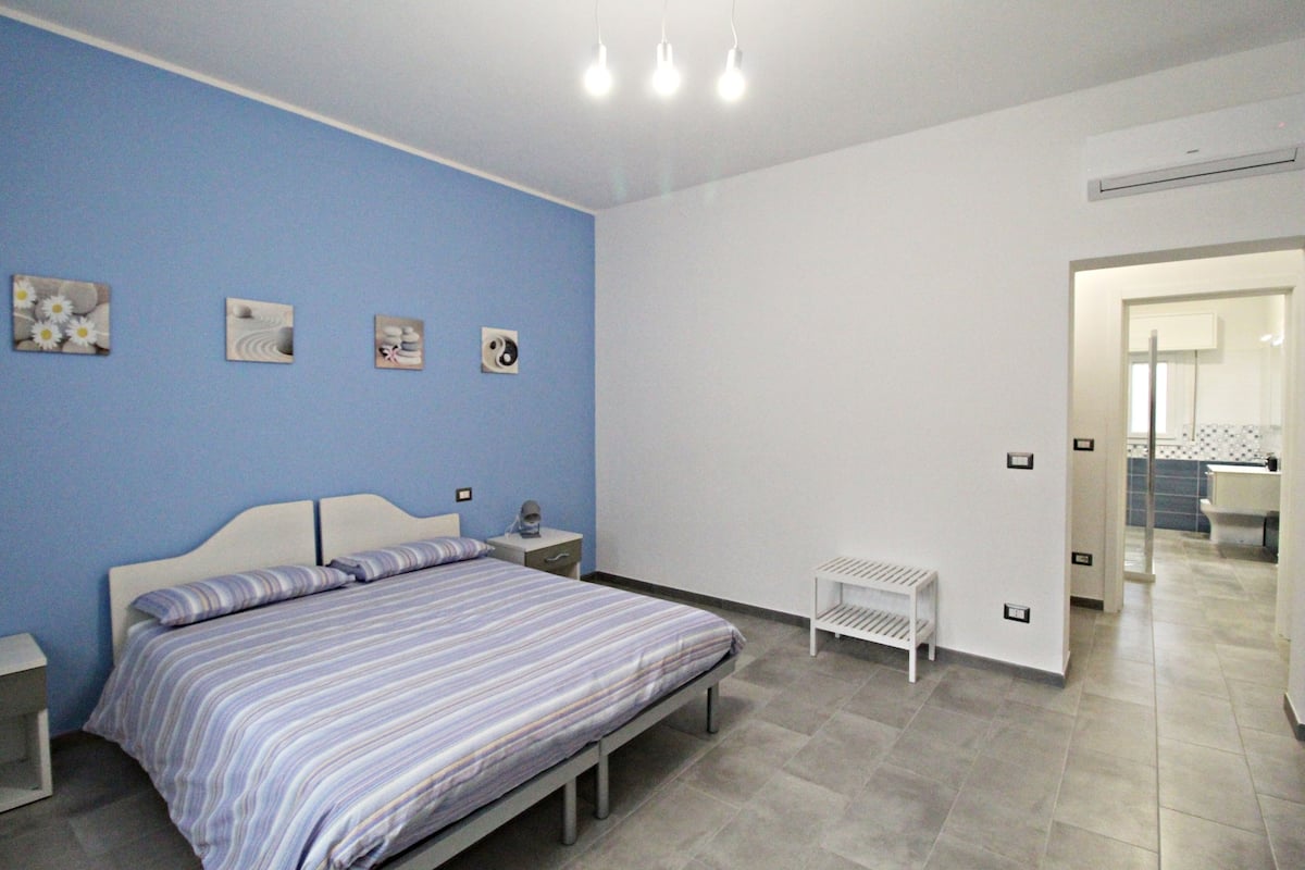 A bedroom with a bed, bedside tables, a small white bench, and a bathroom visible through an open door.