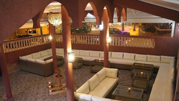 A spacious indoor area with multiple seating arrangements, including sofas and a lounge chair, under a covered section with a tiled floor and a railing. - Maison d'hote Dar amghar (Tafraout)