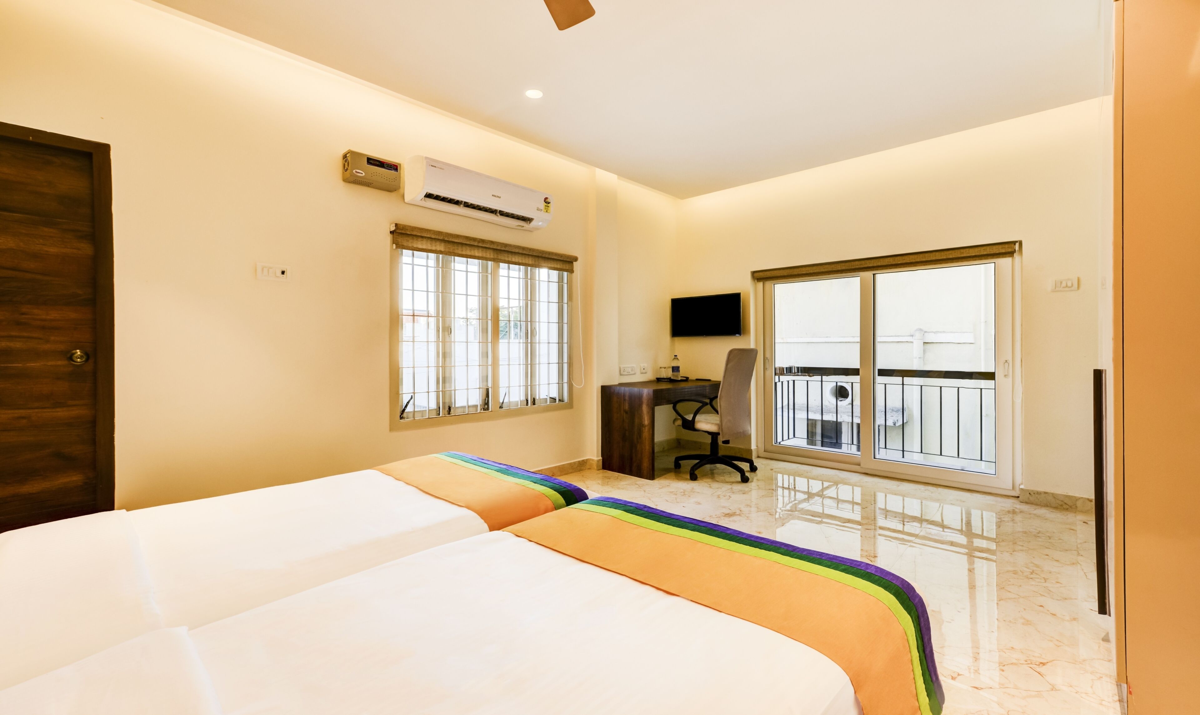 A bedroom with a bed, an air conditioner, a desk with a chair, and a balcony with a railing.