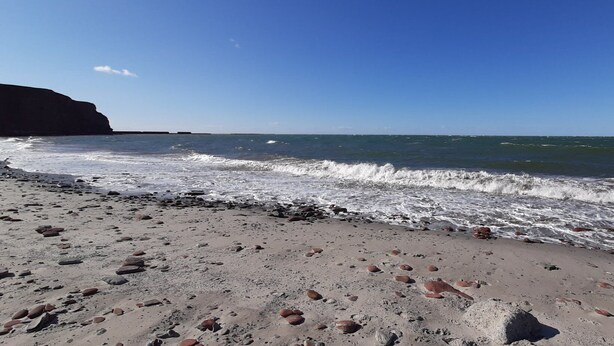 Une plage bordée d’une côte rocheuse, où les vagues se brisent sur le sable, et une falaise en arrière-plan.