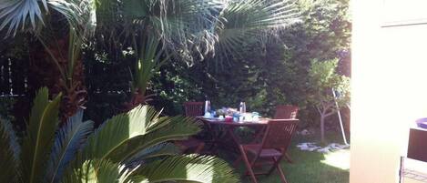 A patio with a table and chairs, surrounded by lush greenery and a palm tree.