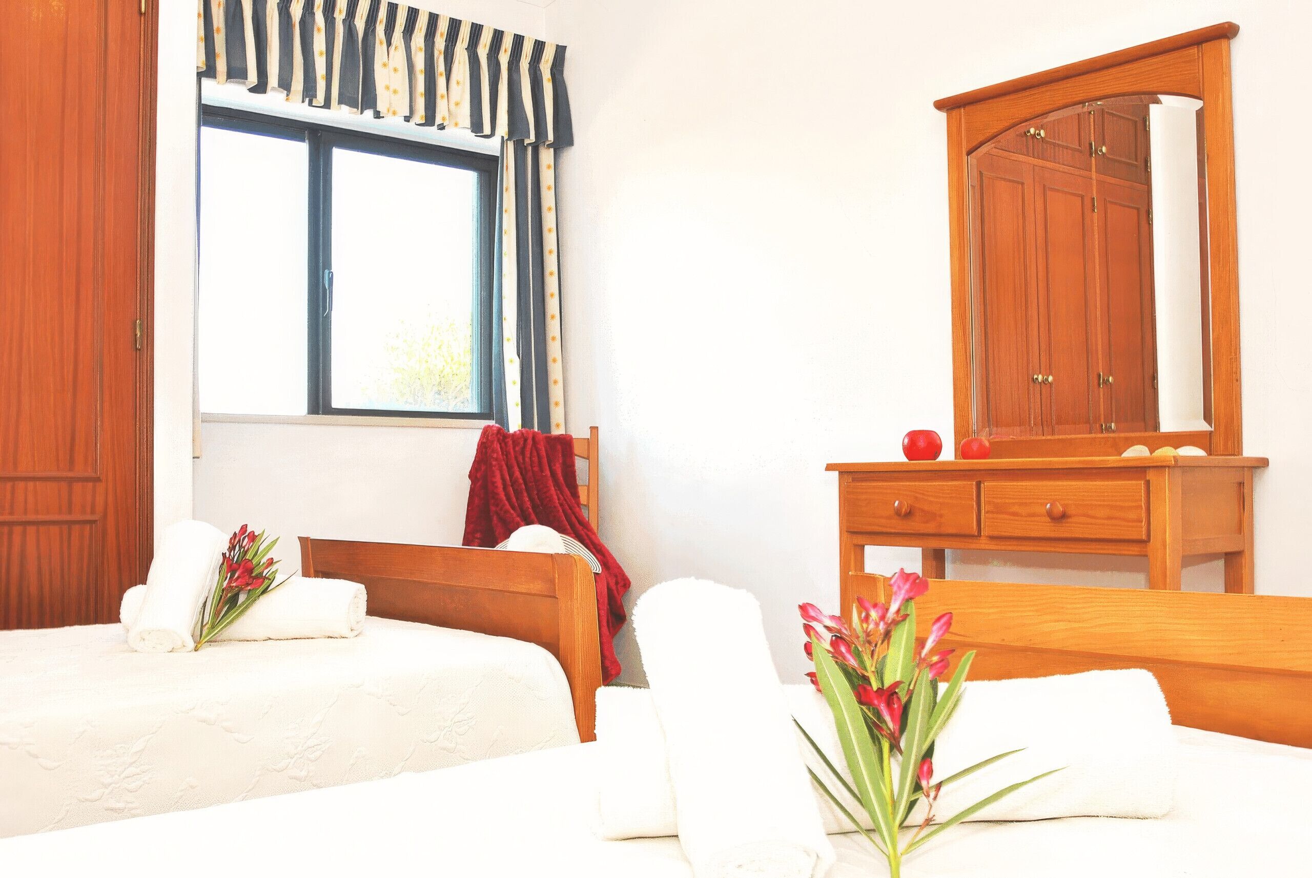 Une chambre d’hôtel avec deux lits, une armoire en bois, une fenêtre avec des rideaux et un vase de fleurs.