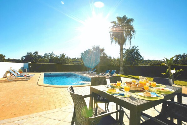 A poolside dining area with a table set for breakfast, lounge chairs, and a swimming pool.