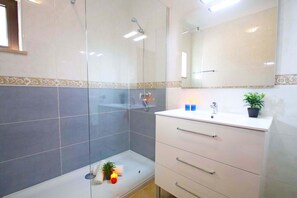 Een badkamer met een glazen douchecabine, een witte wastafel met lades, een spiegel en een plant in een pot op de wastafel.