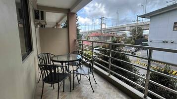A balcony with a table and chairs, a window, and a view of a street with vehicles and buildings.