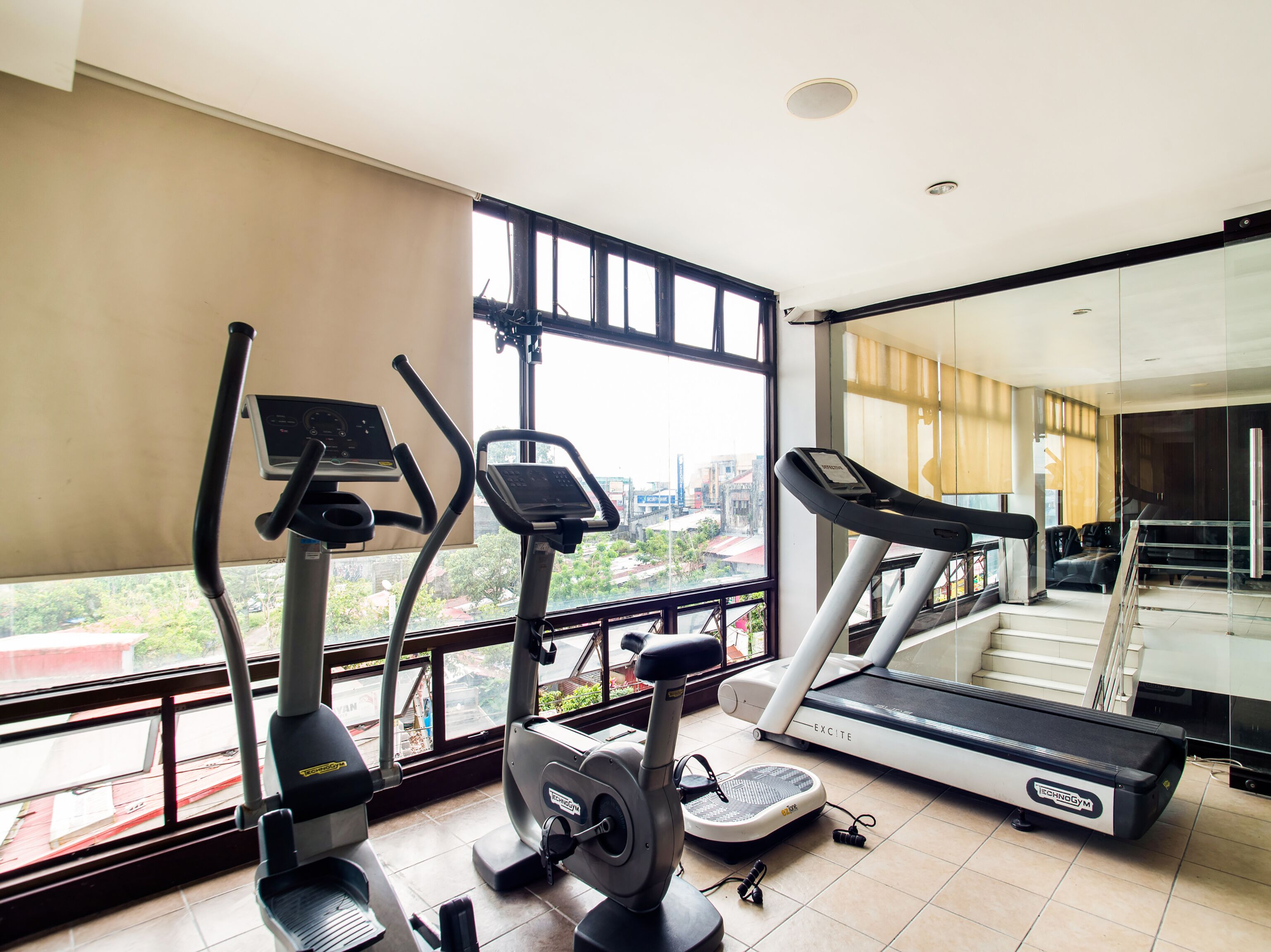 A gym with treadmills, elliptical machines, and a stationary bike, with large windows offering a city view.