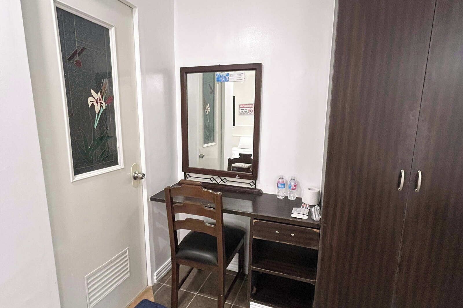A hotel room with a wooden desk, a chair, a mirror, a door with a floral decoration, and a wardrobe.
