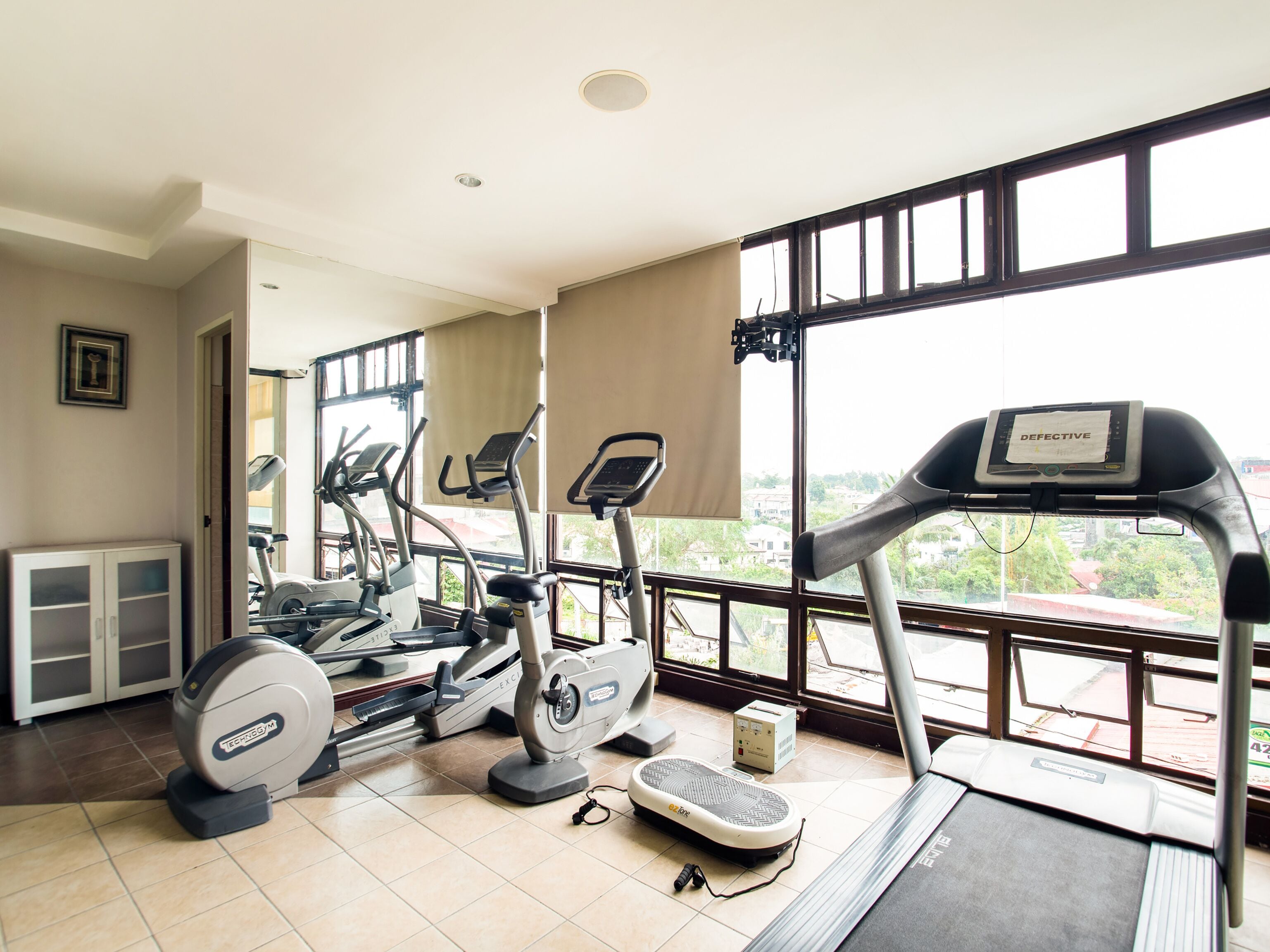 A gym with treadmills, ellipticals, and a rowing machine, large windows with a city view, and a white cabinet.