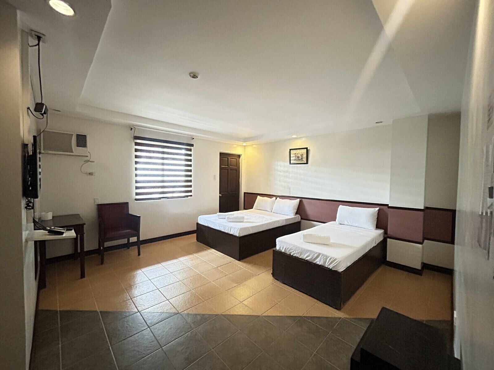 Habitación de hotel con dos camas, un escritorio, una silla, una ventana con persianas y una lámpara en el techo.