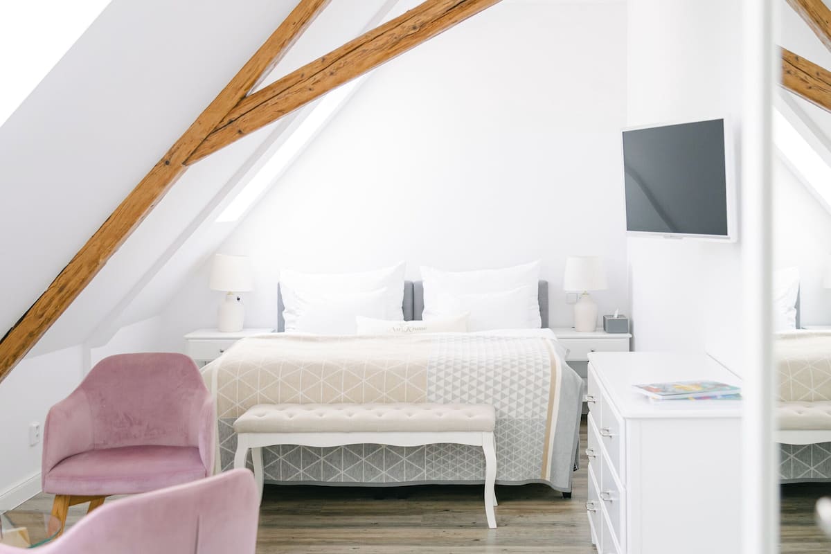 A modern, well-lit attic bedroom with a sloped ceiling, wooden beams, a large bed, a bedside table, a chair, and a TV mounted on the wall.