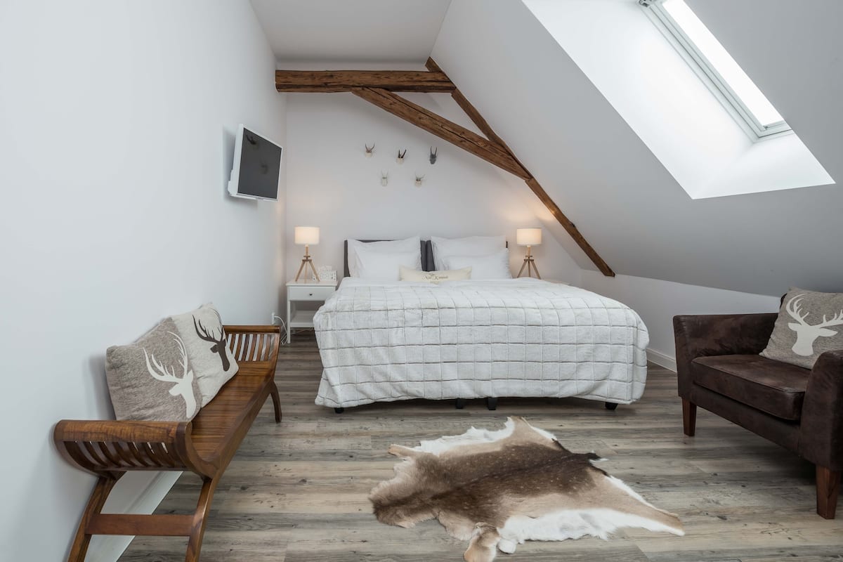 A bedroom with a sloped ceiling, a bed with a white quilt, a wooden bench, a brown sofa, and a fur rug on the floor.