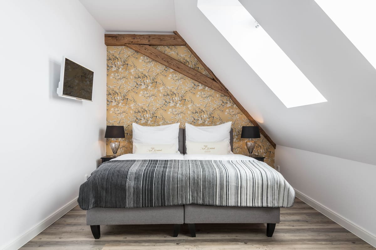 A bedroom with a sloped ceiling, a bed with a striped bedspread, two bedside lamps, and a wall-mounted TV.