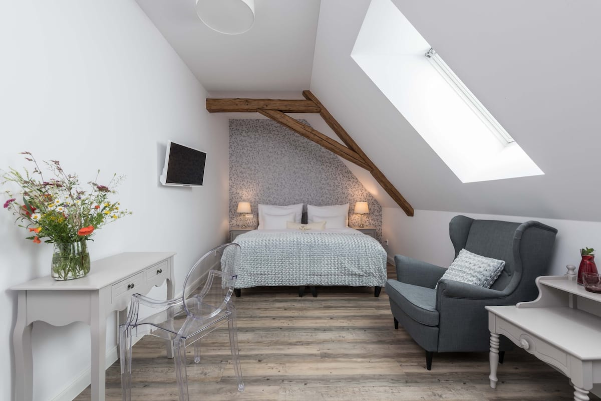 A bedroom with a sloped ceiling, a bed with a patterned bedspread, a grey armchair, a white bedside table, a clear chair, and a vase with flowers.