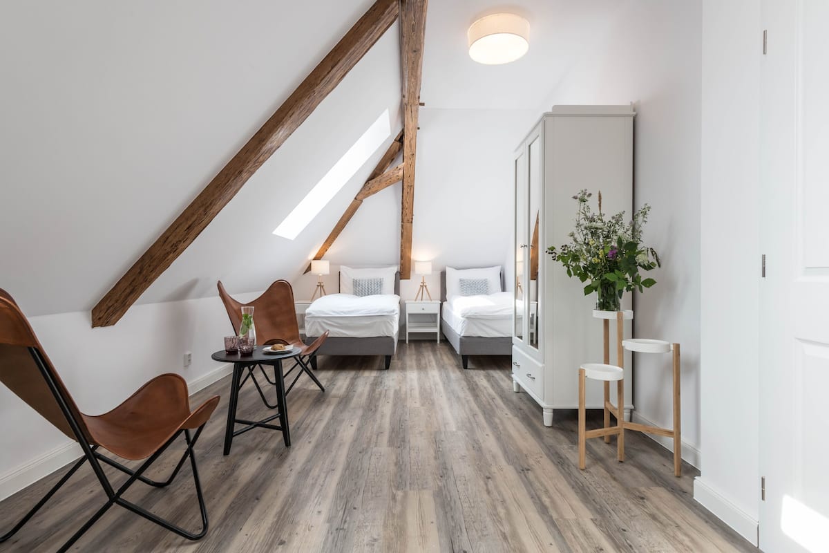 A modern bedroom with a sloped ceiling, wooden beams, two beds, a small table with chairs, a bedside table with lamps, and a potted plant.