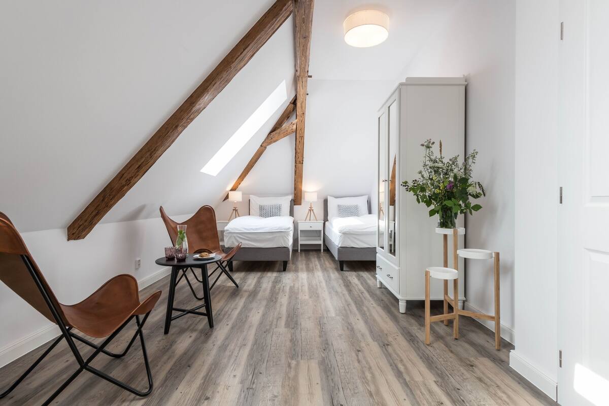 A modern room with a sloped ceiling, wooden beams, two beds, a desk, a chair, bedside tables, and a potted plant.