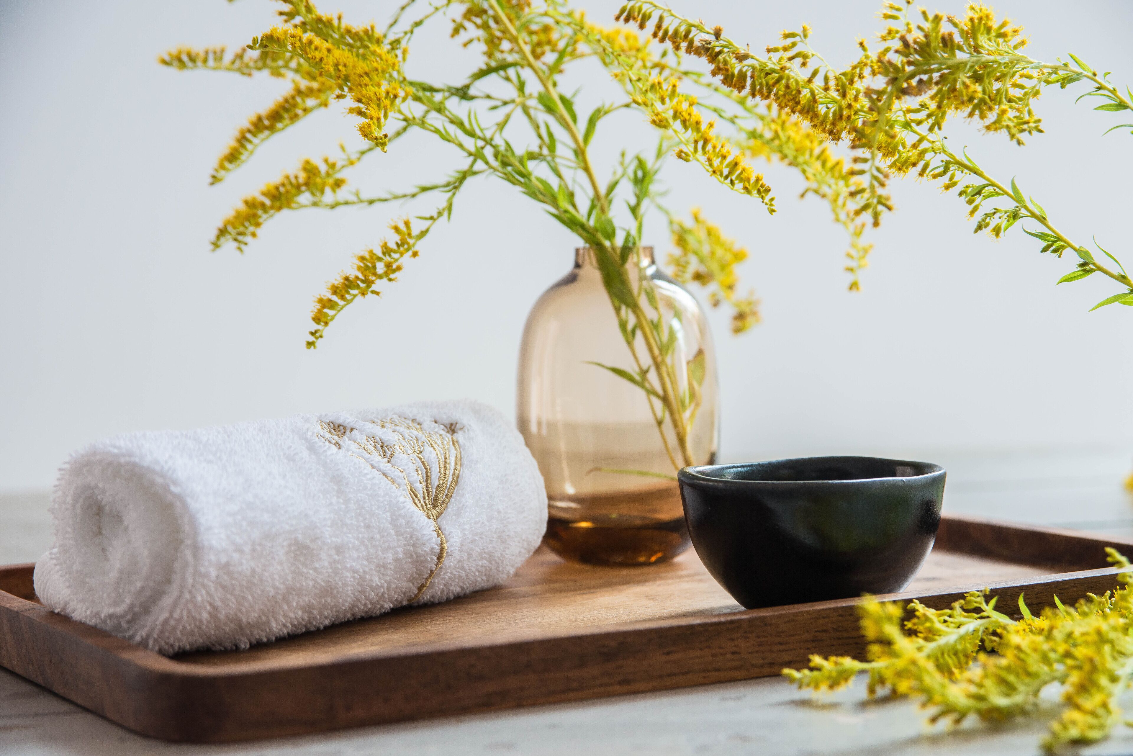 A wooden tray with a rolled white towel, a clear glass vase with yellow flowers, and a black bowl.