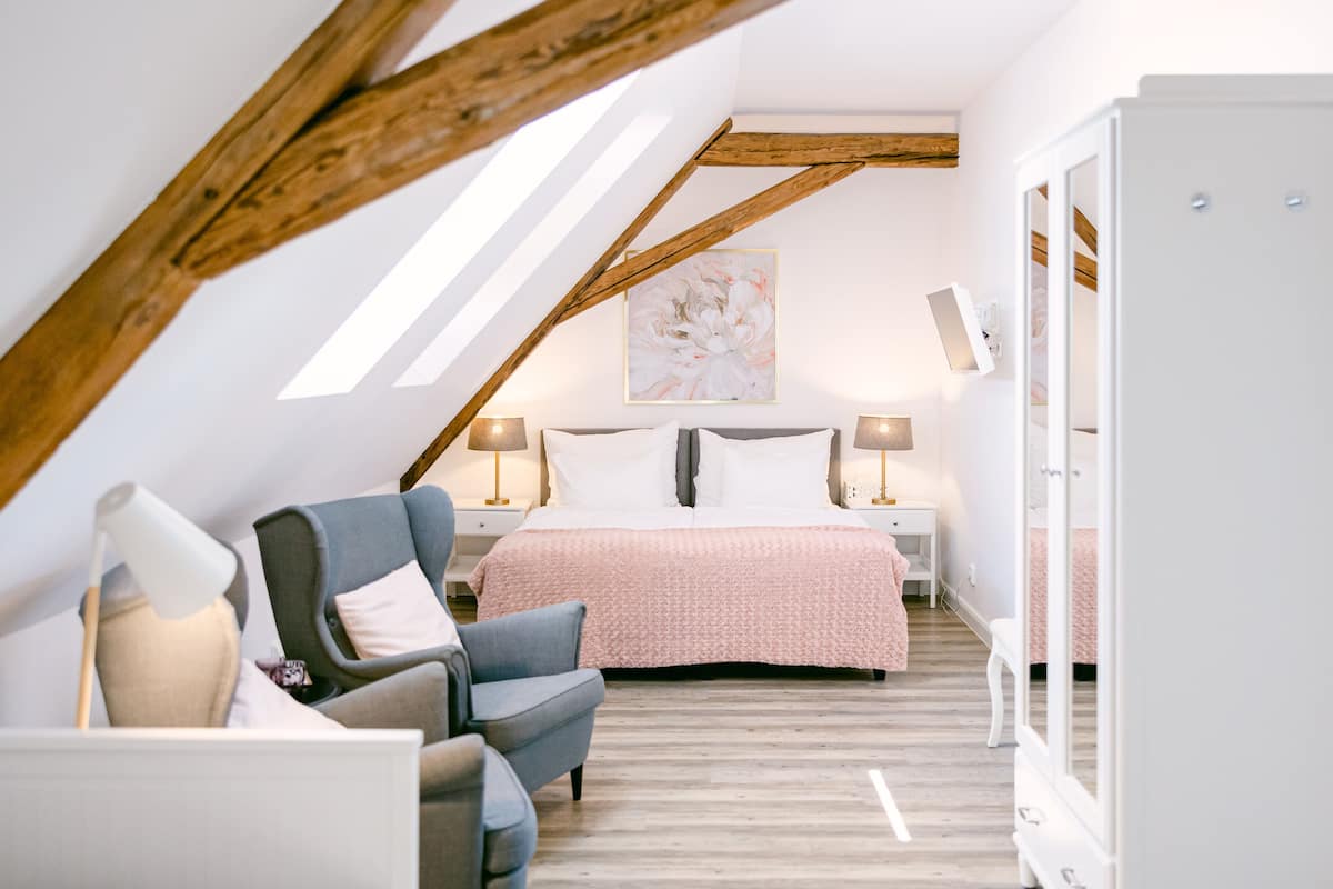 A bedroom with a sloped ceiling, a bed with a pink bedspread, two bedside lamps, a chair, and a large mirror.