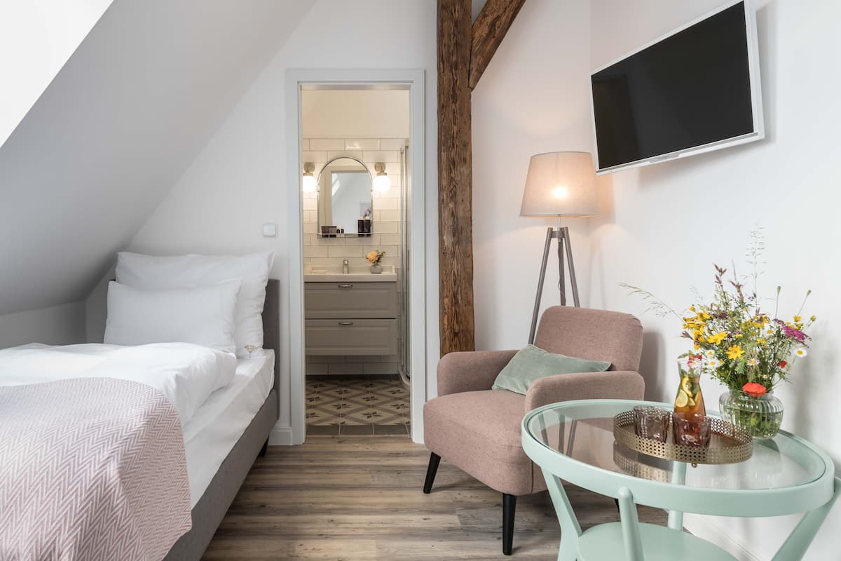 A bedroom with a bed, a chair, a table with drinks, a TV, and a bathroom visible in the background.