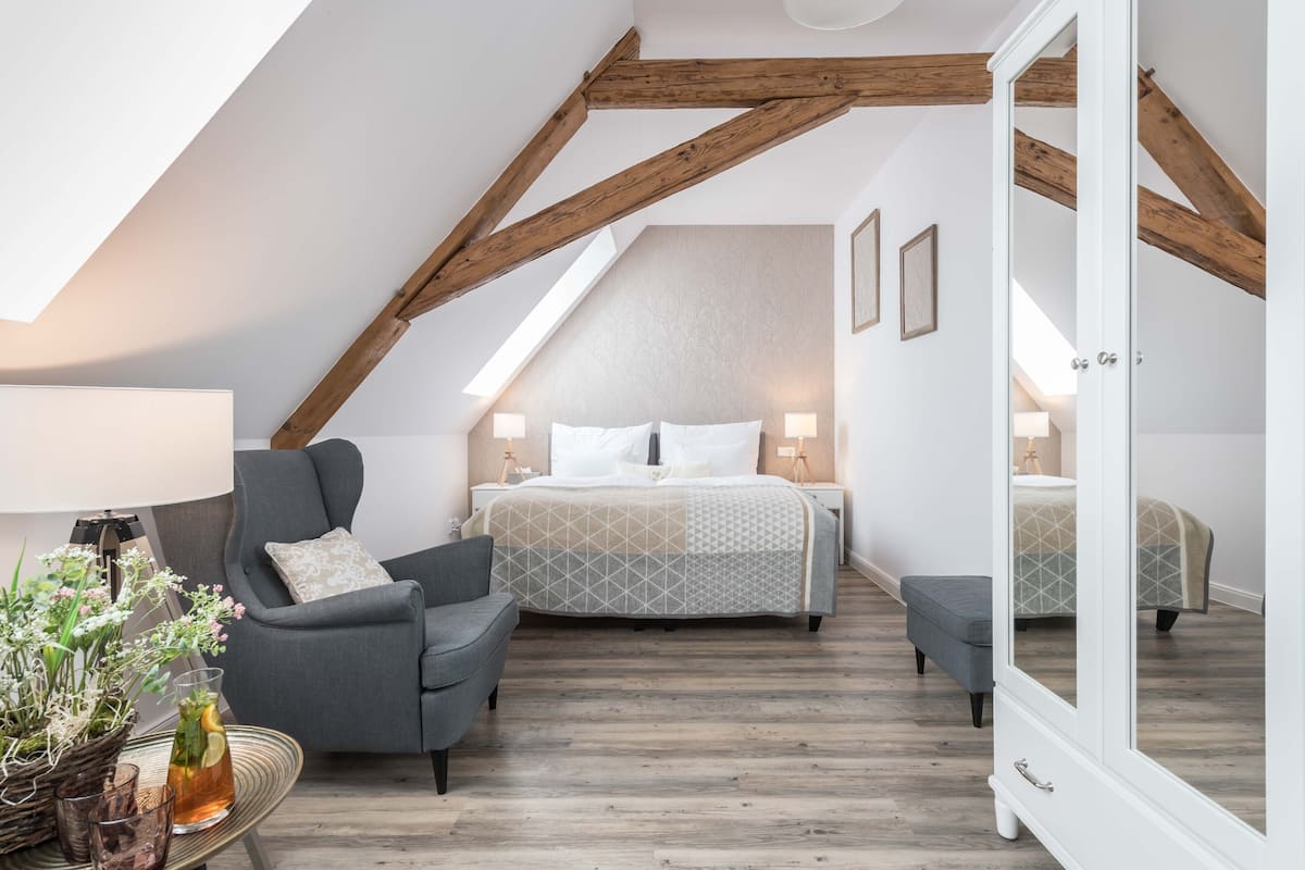 A bedroom with a sloped ceiling, a bed with a patterned bedspread, a gray armchair, a bedside table with a lamp, and a mirror cabinet.