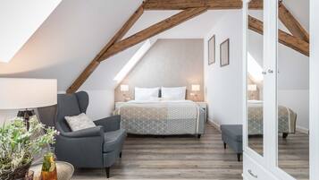 A bedroom with a sloped ceiling, a bed with a patterned bedspread, a gray armchair, a bedside table with a lamp, and a mirror cabinet.