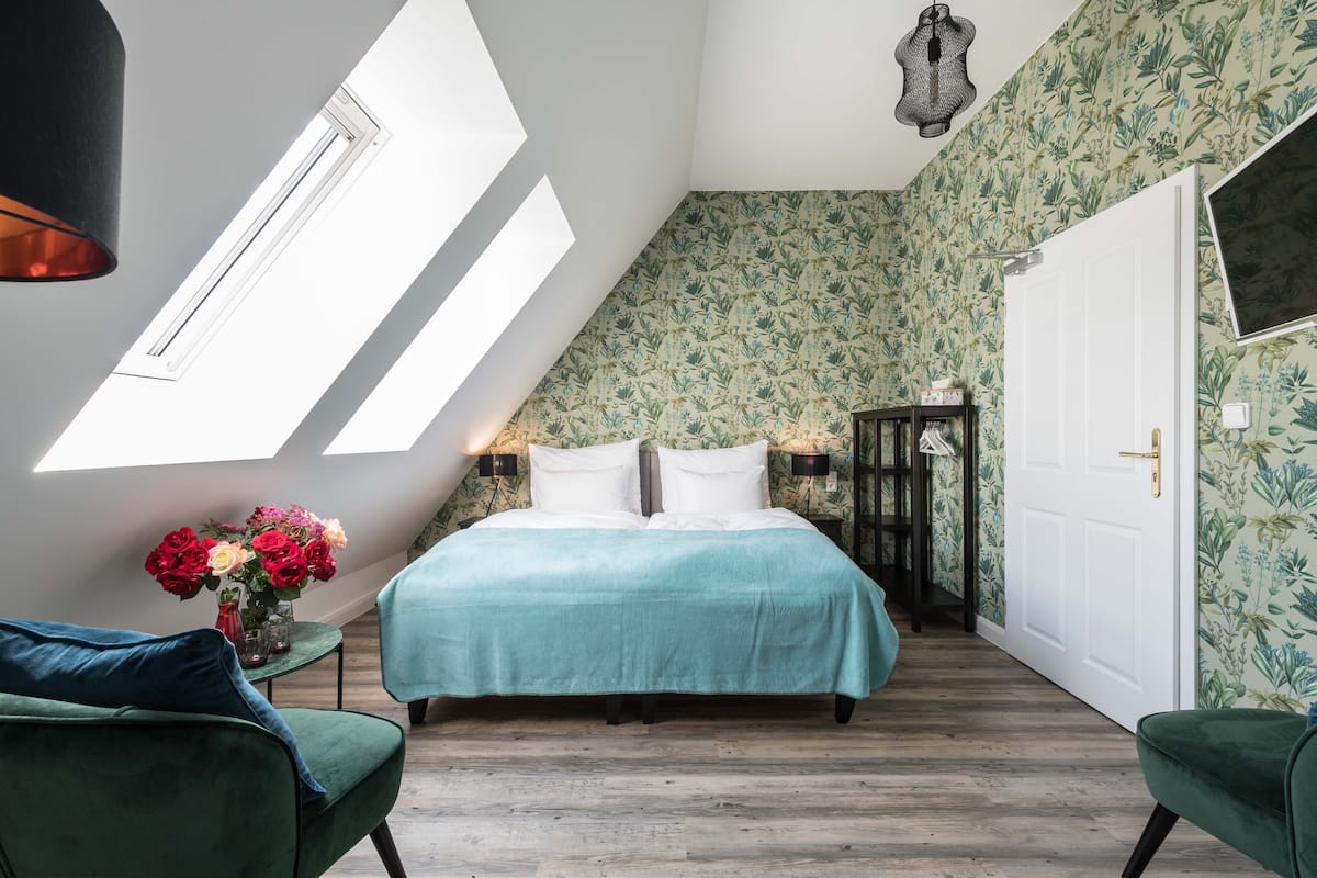A bedroom with a skylight, a bed with a teal bedspread, two green chairs, and a nightstand with a vase of flowers.