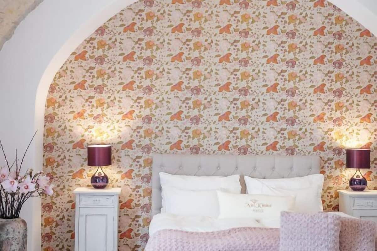 A bedroom with a floral wallpaper, a tufted headboard, a bench, and a chandelier.