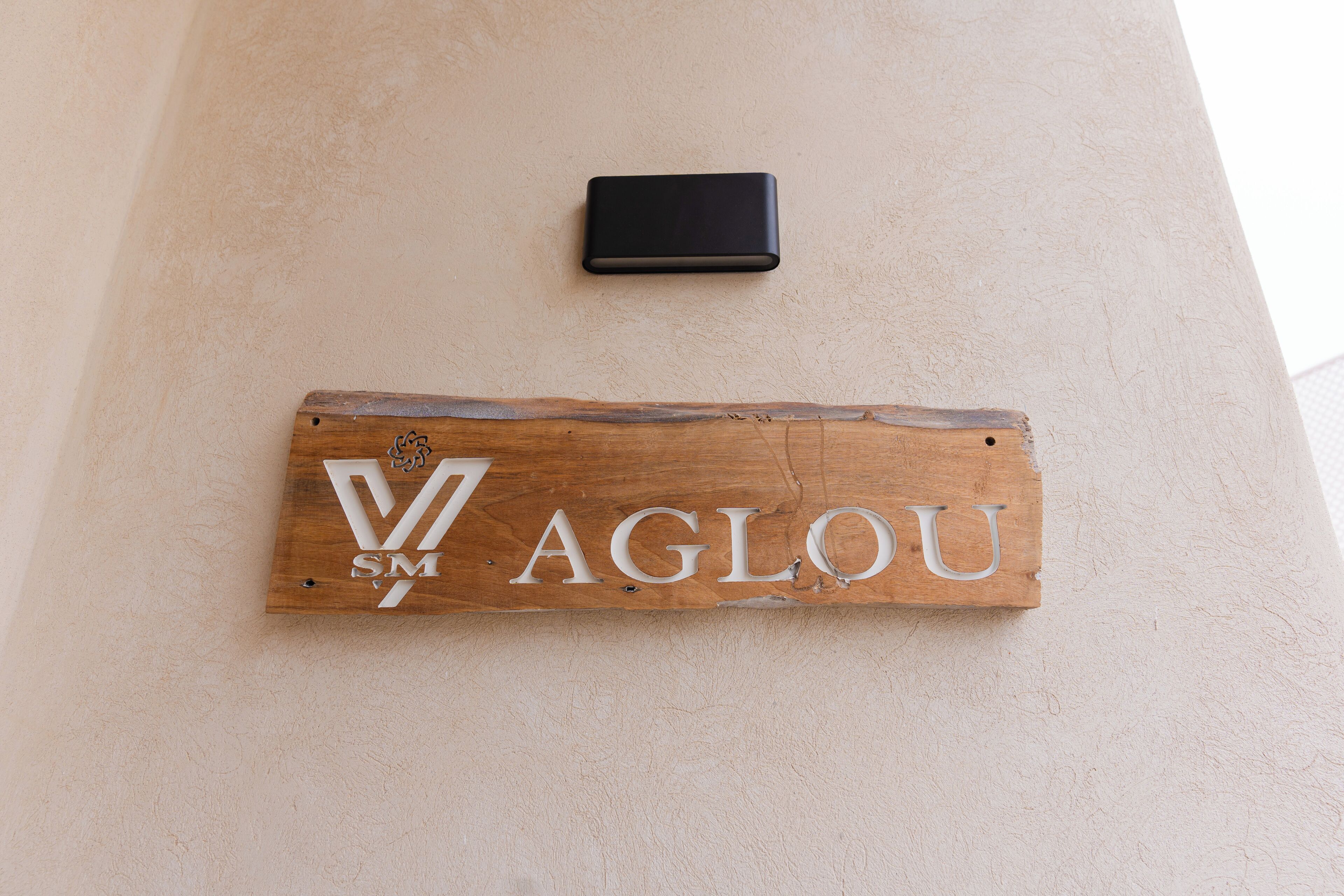Une enseigne en bois portant le nom « AGLOU » et un logo, fixée sur un mur de couleur beige.