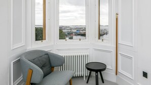 Una habitación con una butaca tapizada de gris, una mesita negra y vistas a un puerto a través de grandes ventanales.
