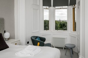Een hotelkamer met een groot bed, een stoel, een kleine tafel en uitzicht op de stad door grote ramen.