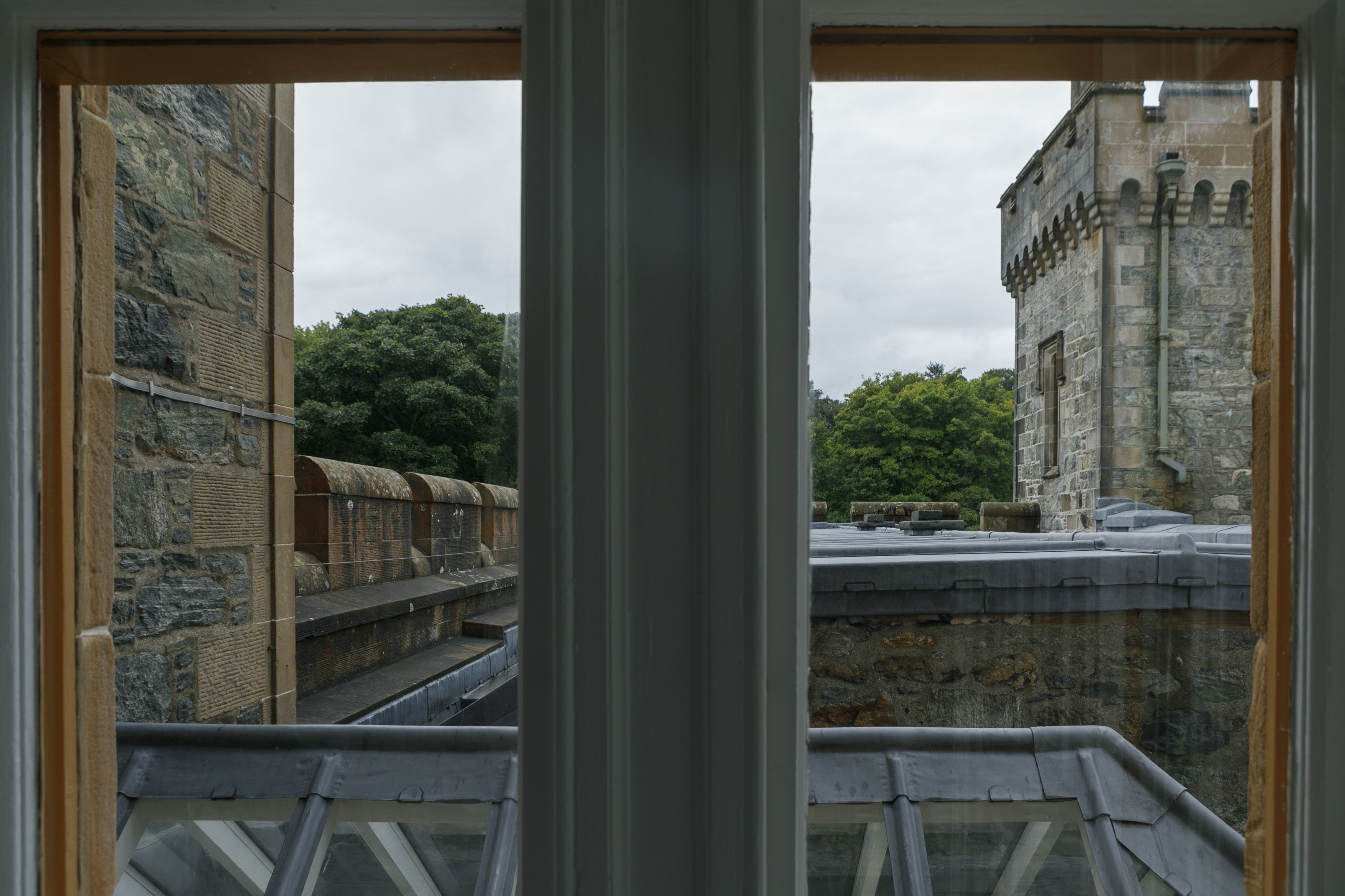 A view from a window overlooking a stone building with a turret, a stone wall, and a green landscape.