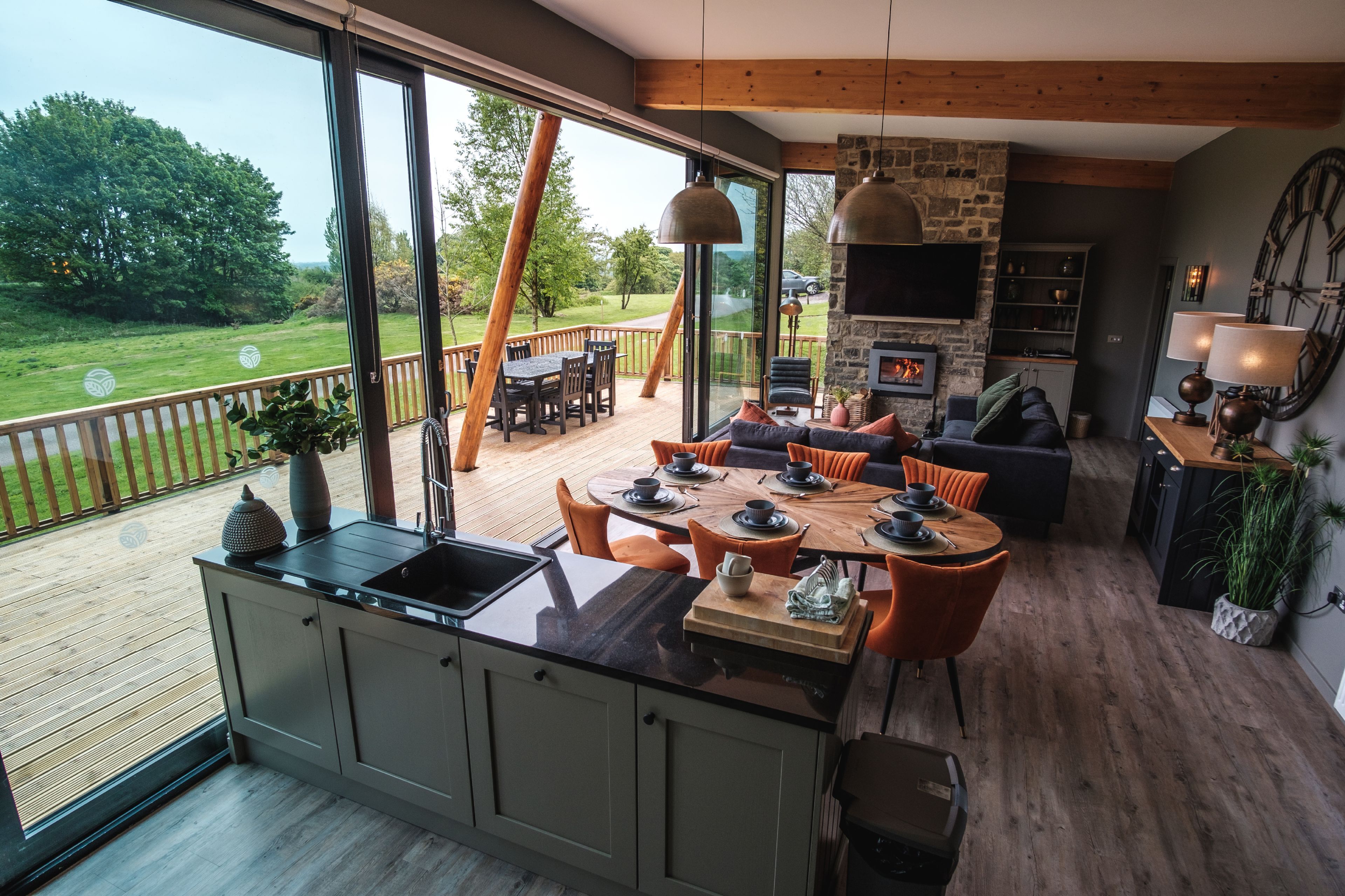 Een ruime woonkamer met een groot raam dat uitkijkt op een terras, een stenen open haard en een eethoek met oranje stoelen.