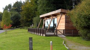 Een moderne houten cabin met glazen ingang, omgeven door weelderige begroeiing en een nummerbord met de locatie.