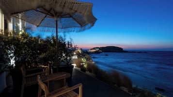 Uma área externa com mesas e cadeiras, um guarda-sol de vime e vista para o oceano ao entardecer.