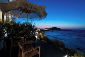 An outdoor seating area with a table and chairs, a wicker umbrella, and a view of the ocean at dusk. - Portara Seaside Luxury Suites (Naxos)