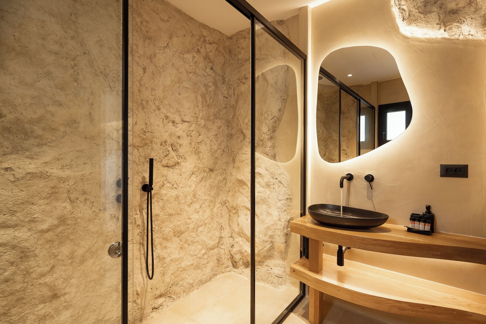 Un baño moderno con pared de textura de piedra, cabina de ducha de vidrio y tocador de madera con lavabo.