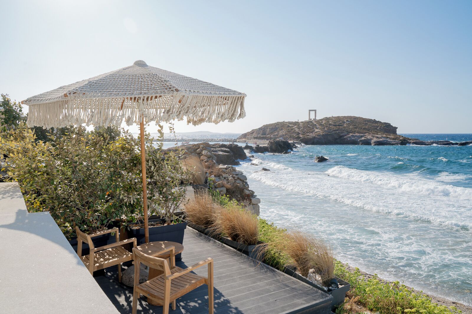 A coastal terrace with a wooden table and chairs, a woven umbrella, and a view of the sea and a rocky outcrop.